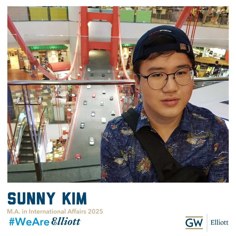 Sunny Kim smiles while sitting down. He is wearing a baseball cap backwards and is wearing a blue button up shirts and blue jeans in front of a bridge model in Thailand. Sunny Kim, M.A. In International Affairs 2025, #WeAreElliott